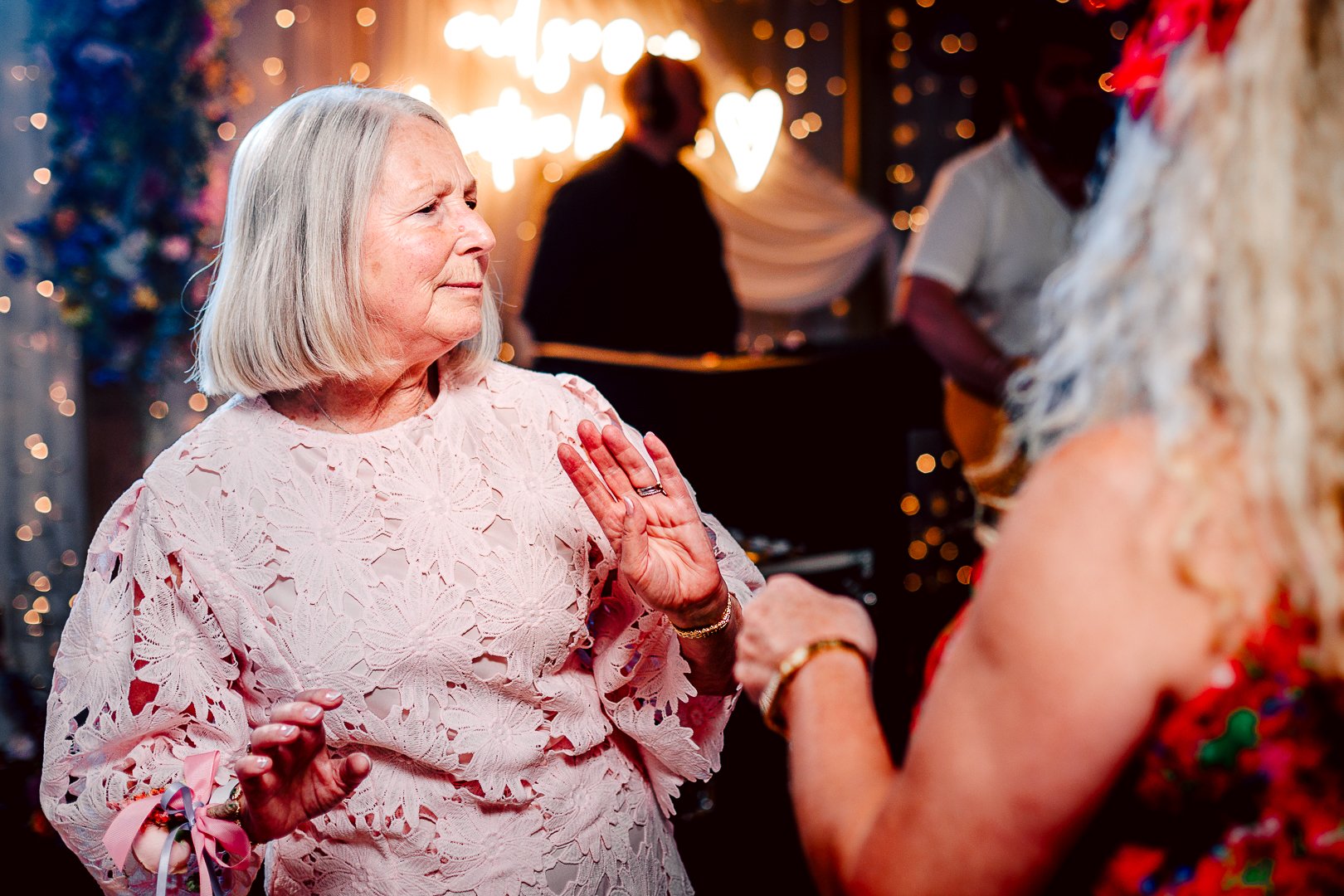 An elderly woman with gray hair wearing a pink lace dress and colorful wrist ribbons is raising her hand in a gesture of oath or celebration while listening to another woman with long blonde hair and a floral dress. The background features warm decor