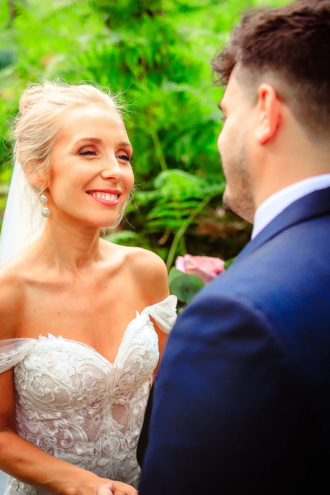 A bride and groom are exchanging vows outdoors in a lush green setting. The bride is smiling and wearing a white off-the-shoulder lace wedding dress with pearl earrings. The groom, in a dark suit, faces her with his back to the camera.