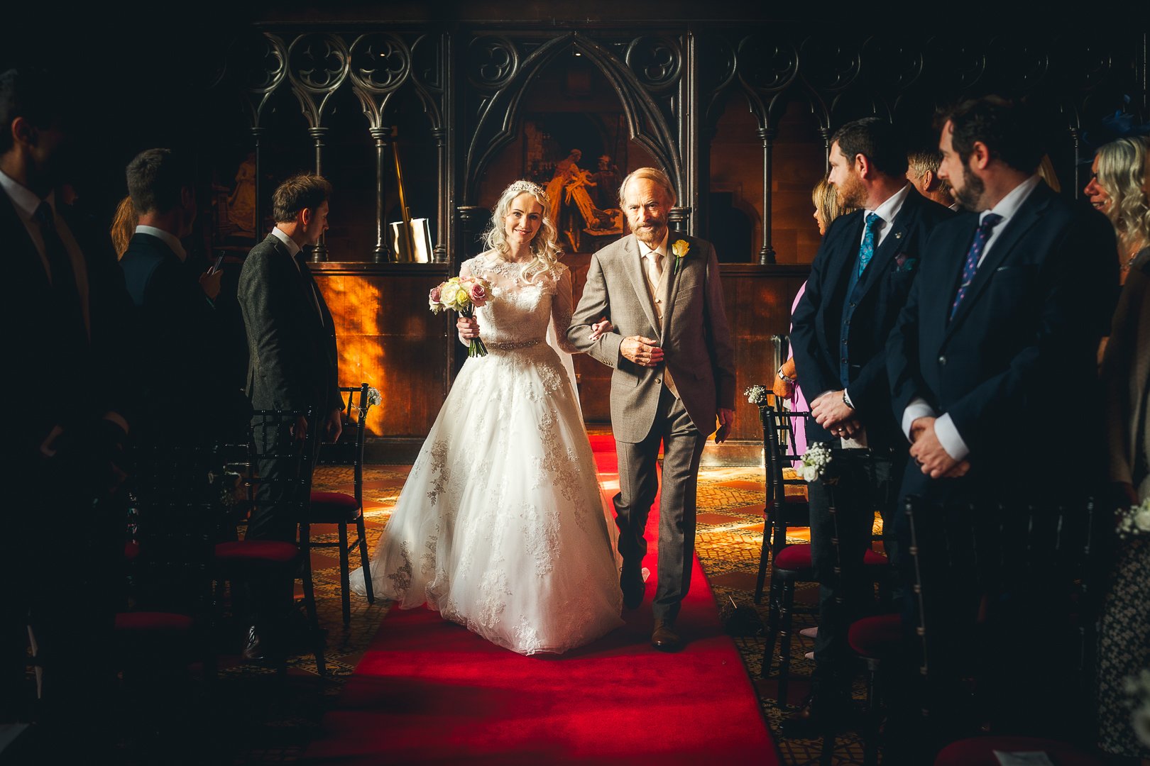 Bride walking down the aisle with her father during a wedding ceremony in a dimly lit church. The bride is holding a bouquet and wearing a white wedding gown, while the father is dressed in a light-colored suit. Guests are standing on either side, wa