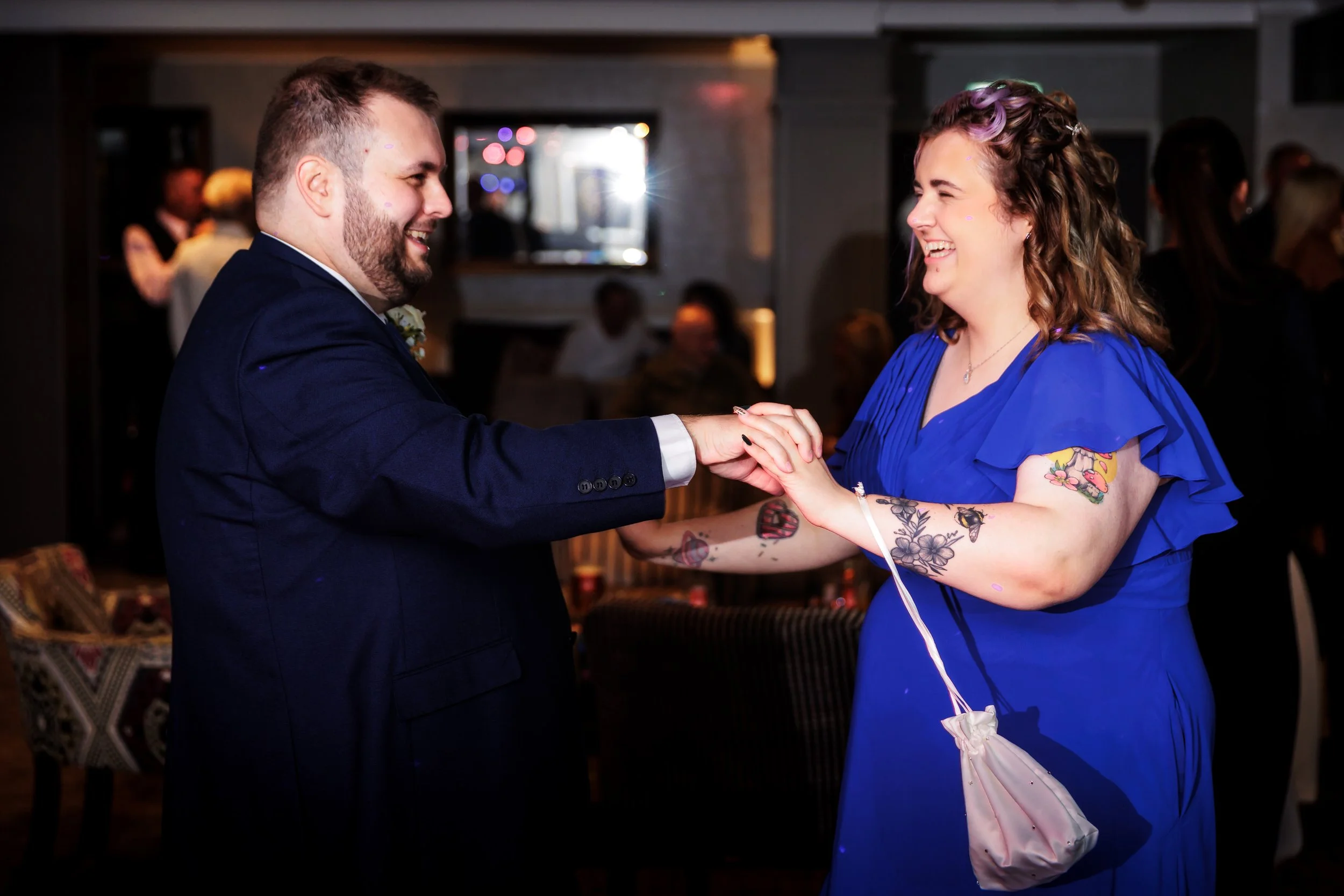 A man and woman dance together at a celebration, smiling and holding hands, in a dimly lit room with other guests in the background.