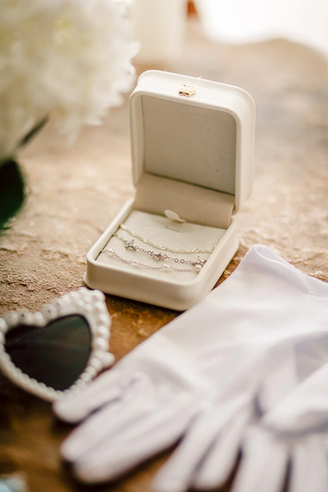 Open jewelry box containing silver bracelets on a textured surface next to white gloves, a heart-shaped mirror, and a white cloth.