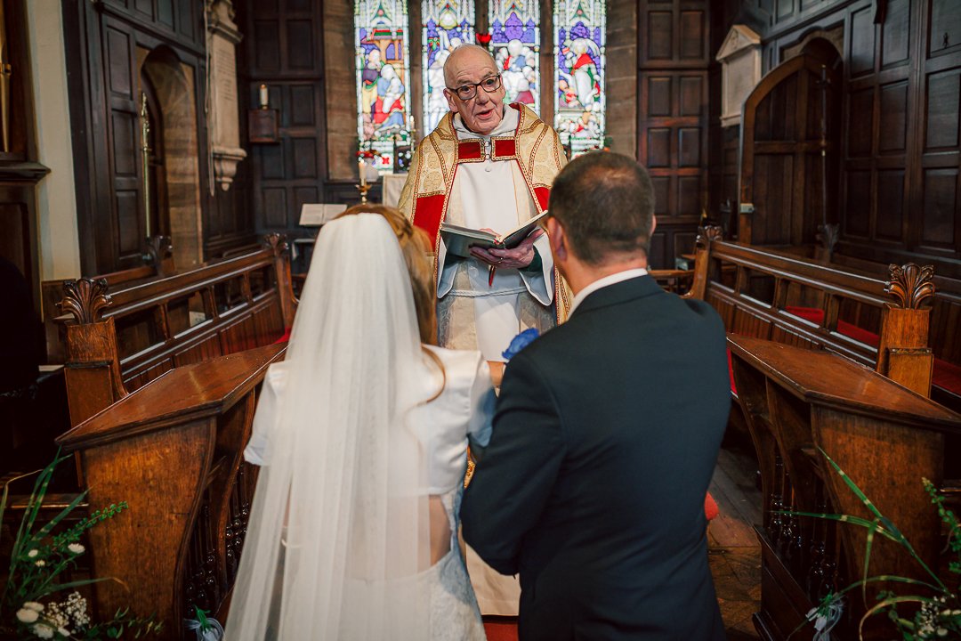 Bride and groom hold hands as they say their vows during their wedding ceremony at Saint Martins Church in Sale. The vicar looks on as the three stand at the alter in front of the large pane glass window.