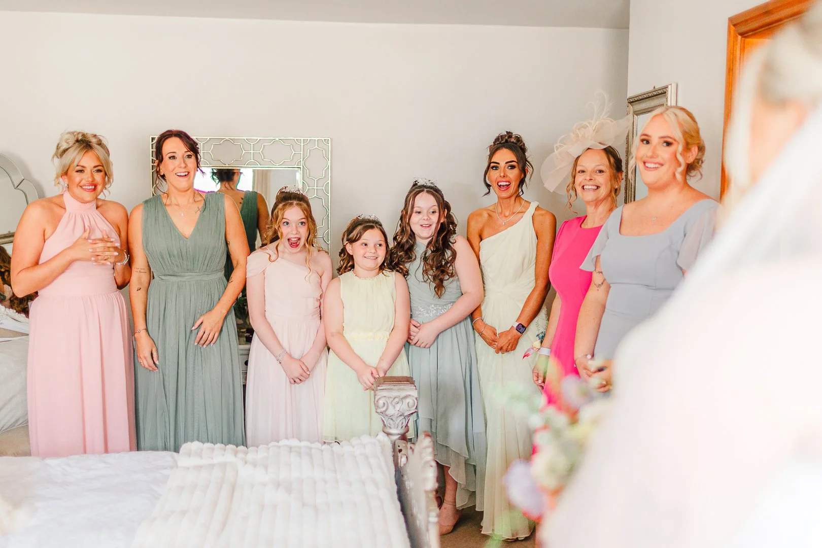 Bridemaids and flower girls in pastel dresses smiling and looking at a bride in a white veil, in a bedroom with a mirror and a bed during wedding day perperations at Beeston Manor in Preston