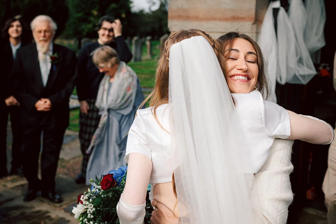 Hugs for the bride as a guest embraces Zoe the bride on her wedding day surrounded by family members in the grounds of Saint Martins Church in Sale