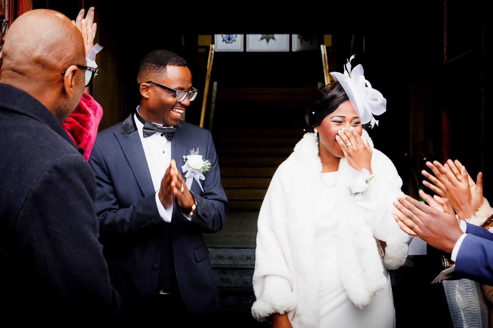 A bride in a white dress and fur coat is laughing with her hand covering her mouth while being clapped for by a group of people at her wedding.