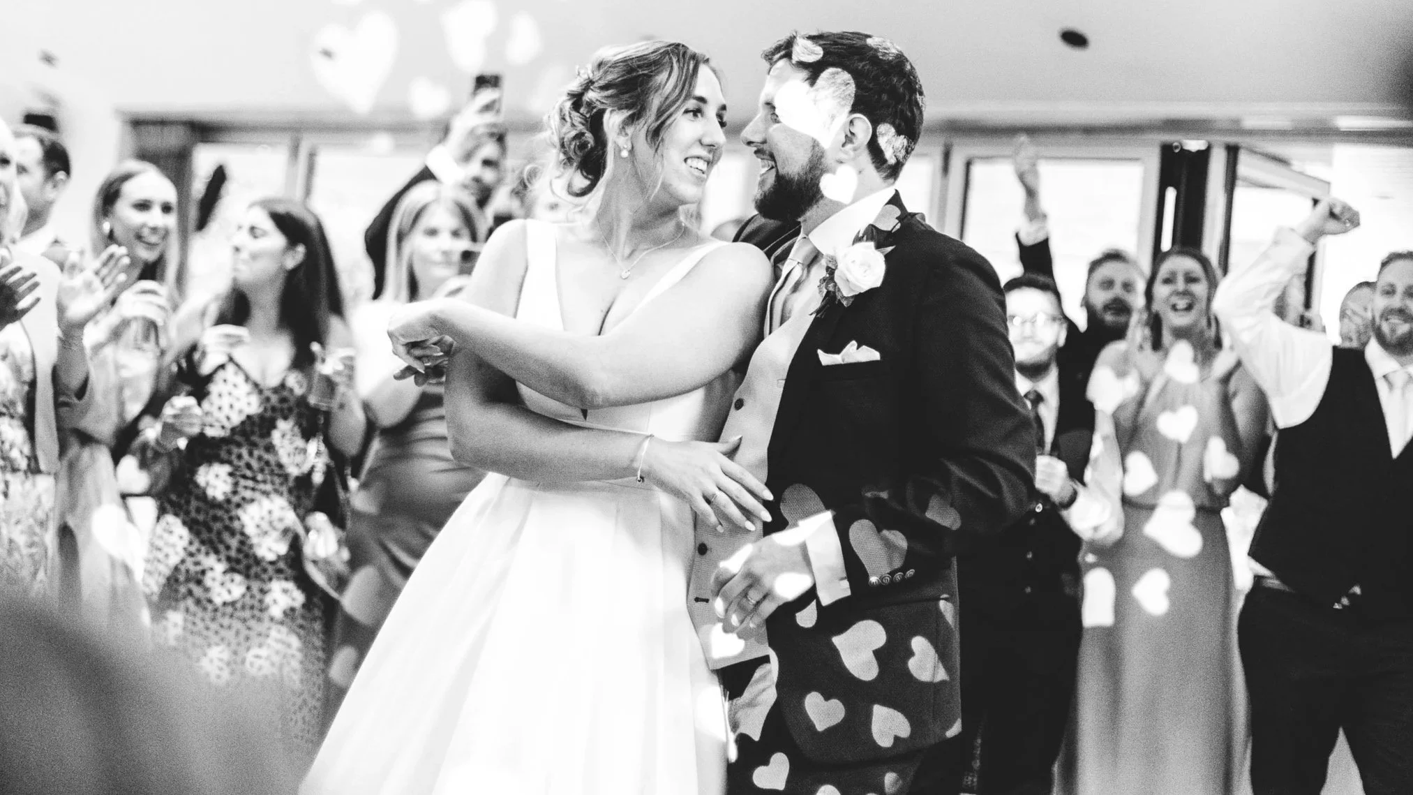 Bride and groom surrounded by guests and heart-shaped confetti during their wedding reception.