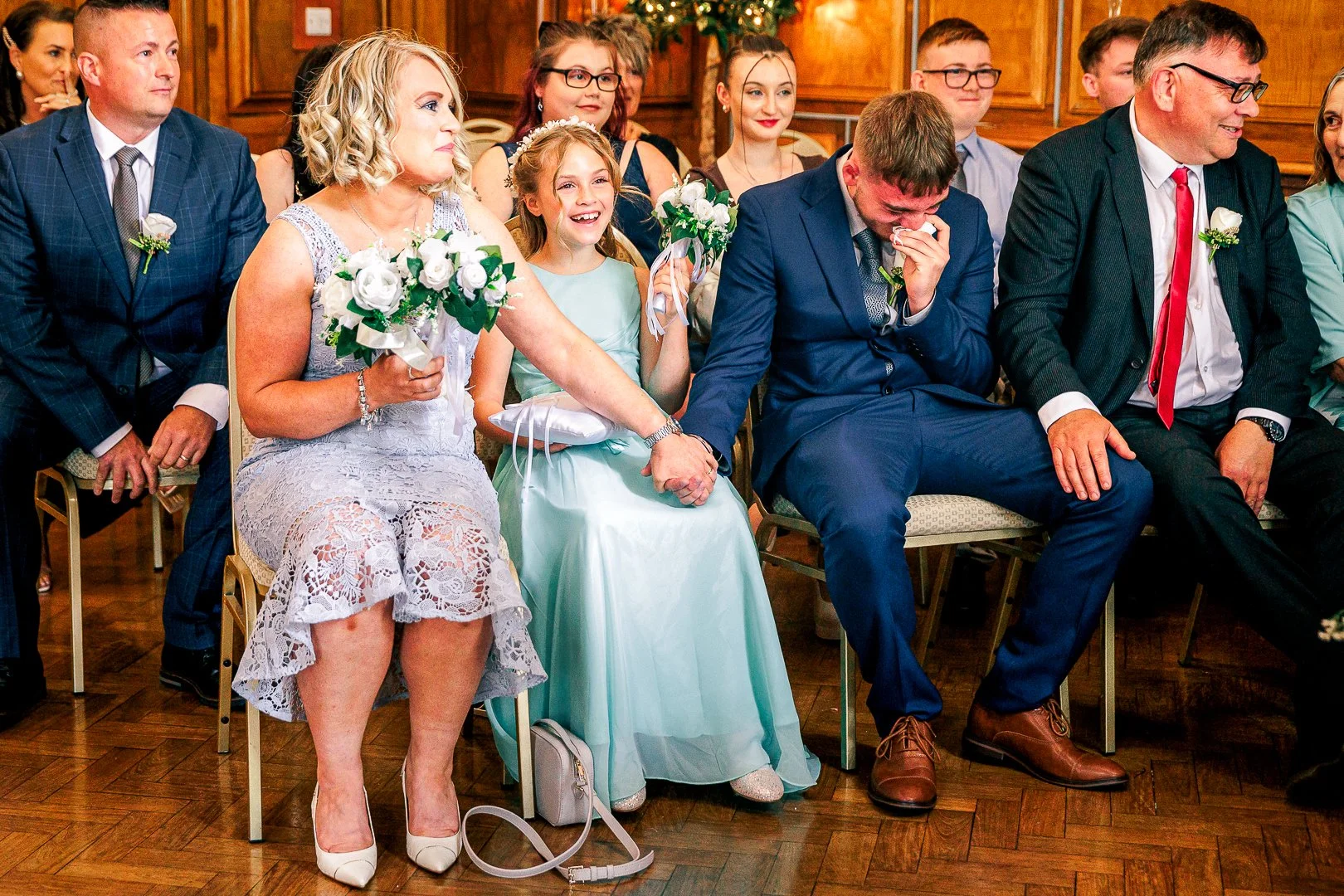 People sitting in a row at a wedding, including a woman in a lace dress, a girl in a light blue dress, a man in a blue suit, and a man in a black suit with a red tie, all holding hands or touching each other, with joyous expressions.