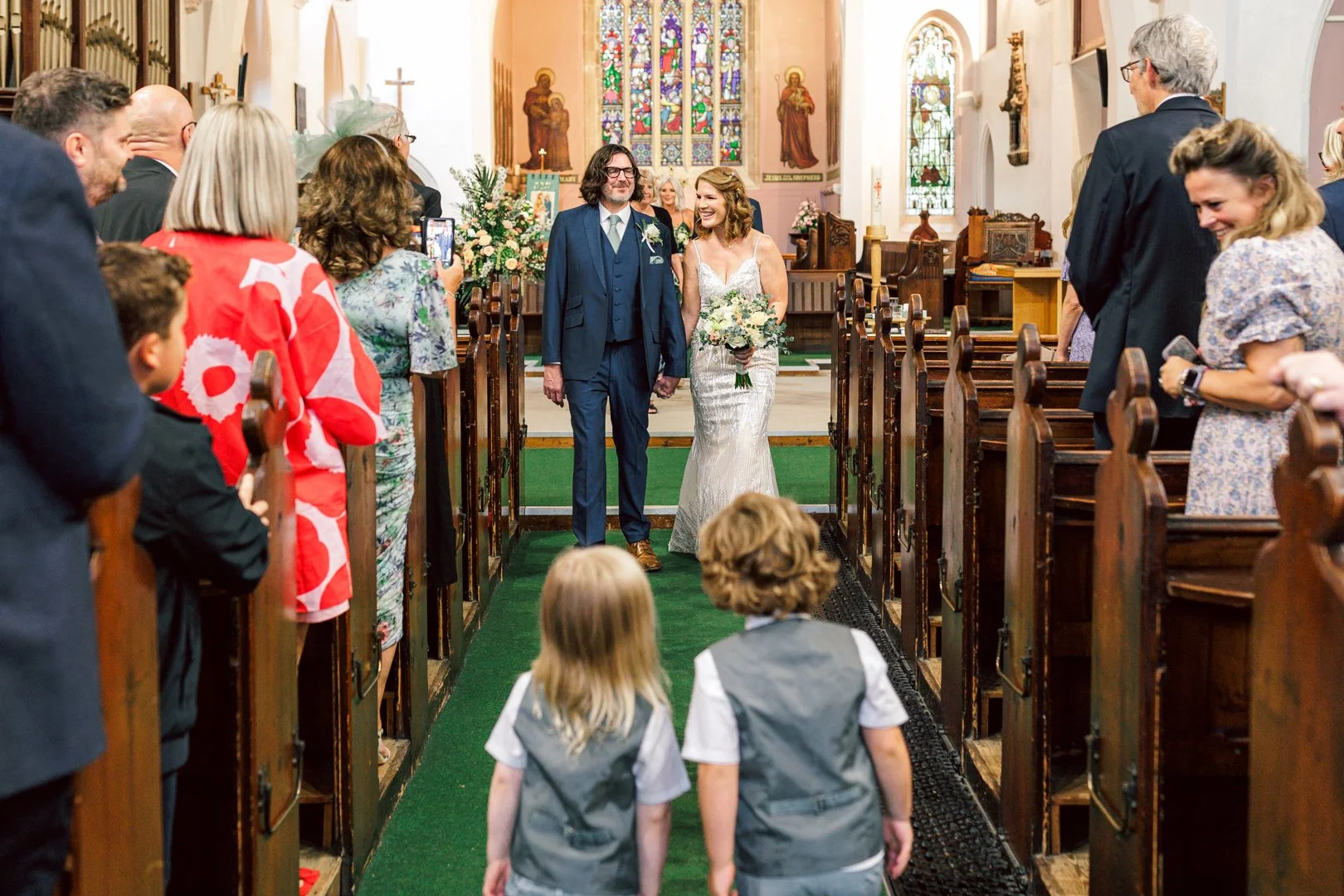 A bride and groom walking down the aisle inside a church, holding hands, smiling at each other. Guests are on either side, some taking photos, celebrating the wedding.