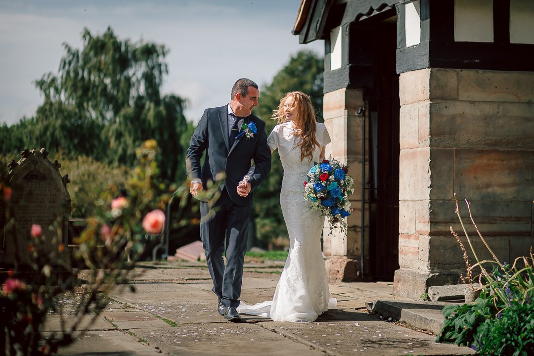Zoe and Sam walk out of Saint Martins Church in Sale following their wedding, Zoe holding a blue floral bouquet matching the grooms blue buttonhole