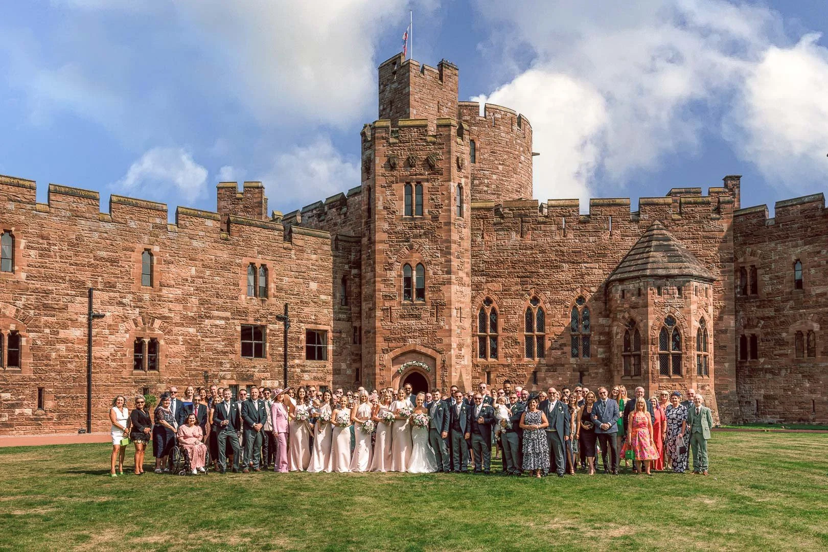 Weding guests join the bride and groom for a full wedding party photo in front of Peckforton Castle in Cheshire