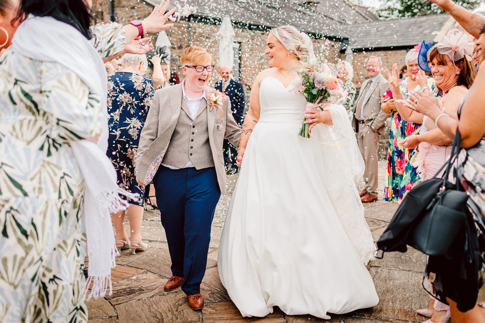 Joyful confetti moment for 2 brides walking happily through a their cheering guests who are throwing confetti following their wedding ceremony at Beeston Manor in Preston