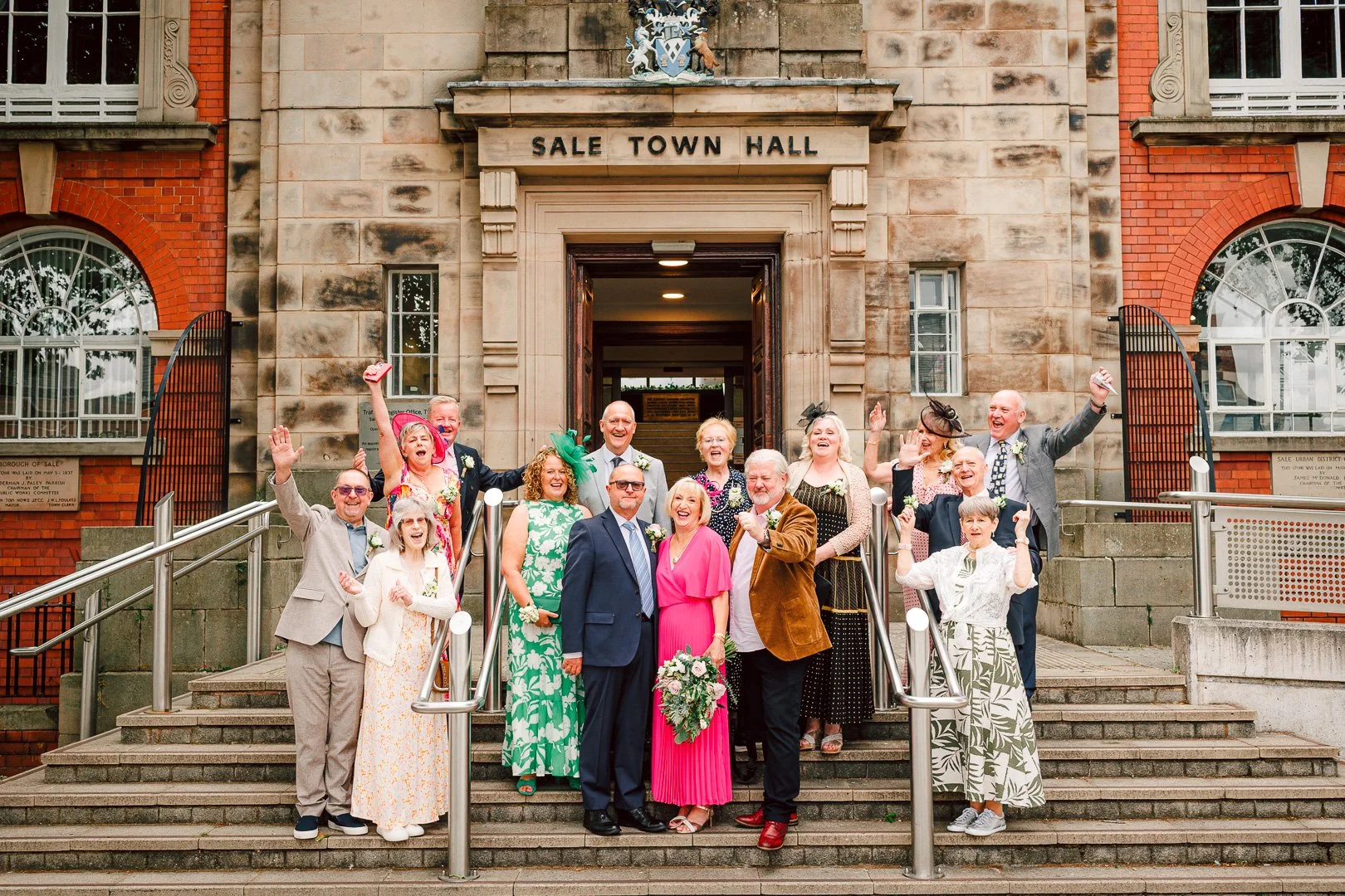 Full wedding party celebrating on the exterior stone steps of the building, captured by a local wedding photographer at Sale Town Hall.