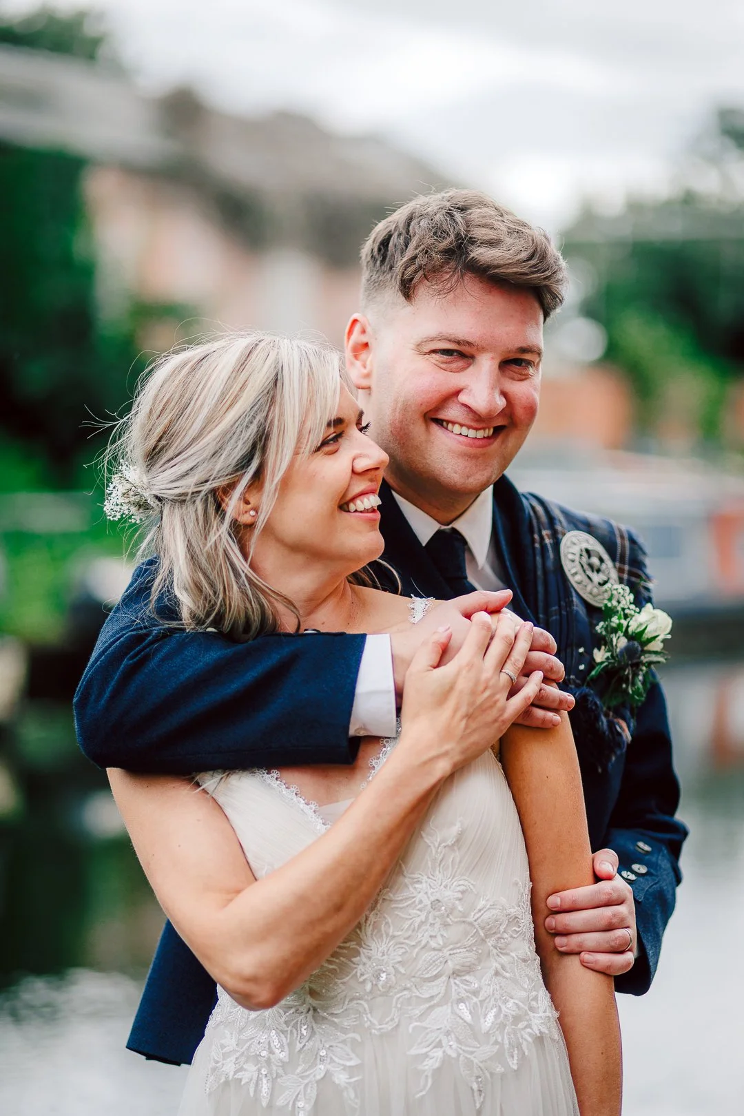 A newlywed couple, with the groom in a dark suit and the bride in a white lace wedding dress, sharing a joyful moment outdoors during the daytime.