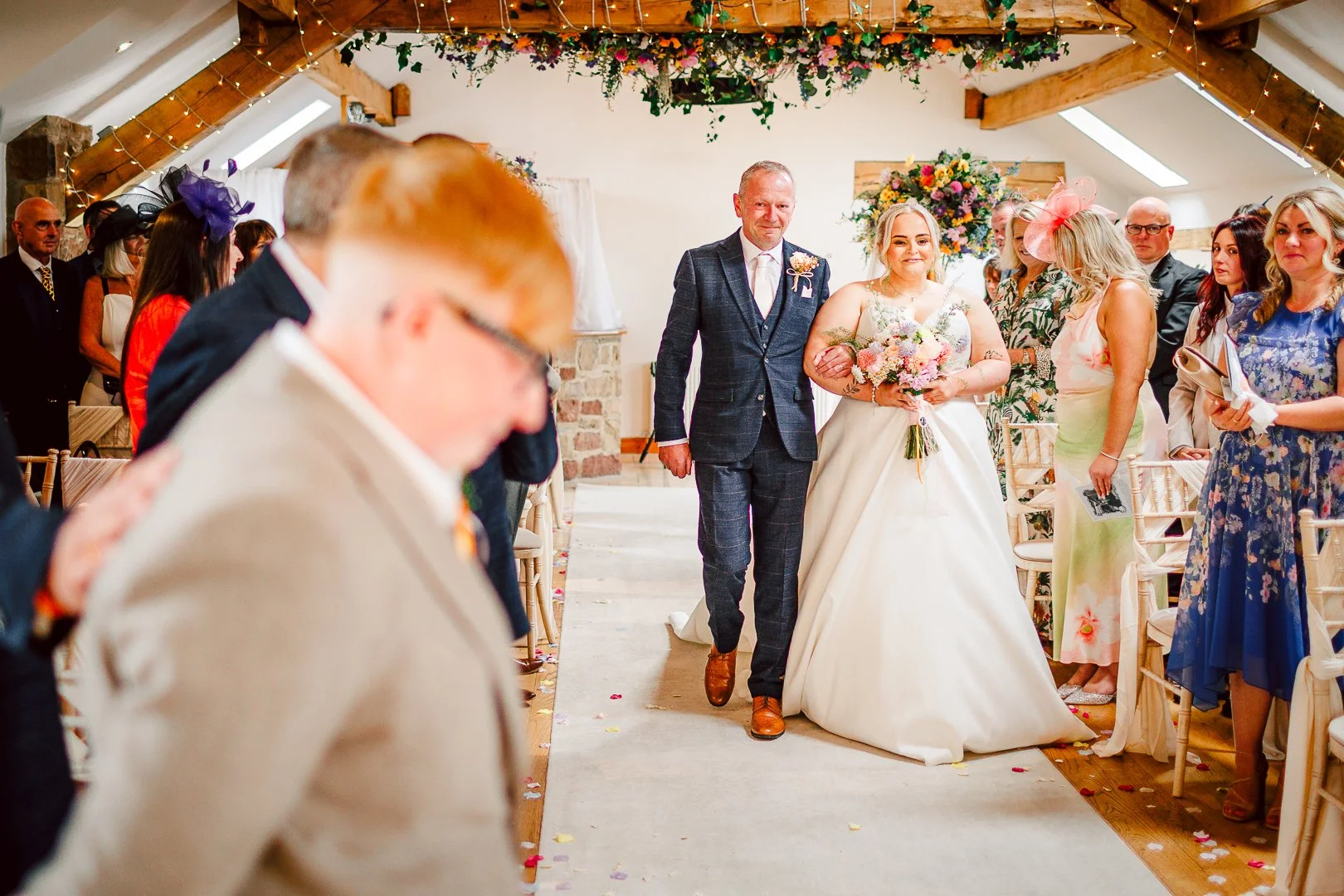A bride walking down the aisle in a wedding ceremony, flanked by a man, likely her father, in a suit. Guests are seated on either side, some standing and watching as she makes her way. The venue is decorated with flowers and string lights, with a woo