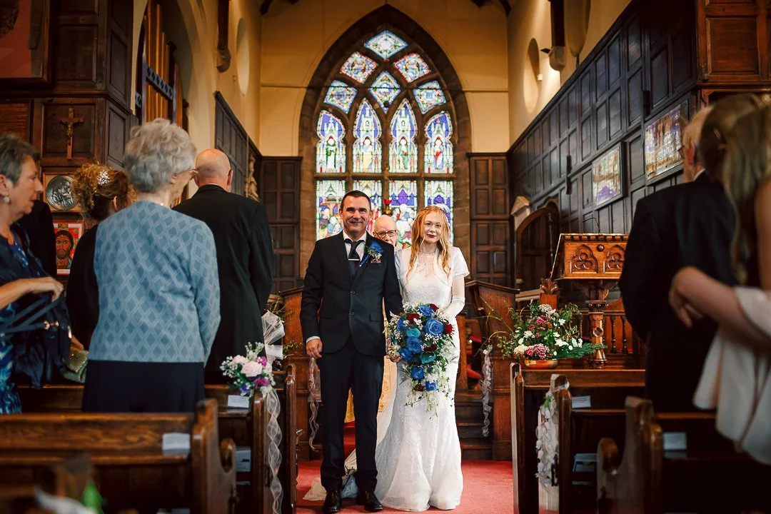 Groom dressed in dark blue suit with a blue buttonhole and bride dressed in white complete with blue and red floral bouquet standing at the front of Saint Martins Church in Sale surrounded by guests in front of pane glass window