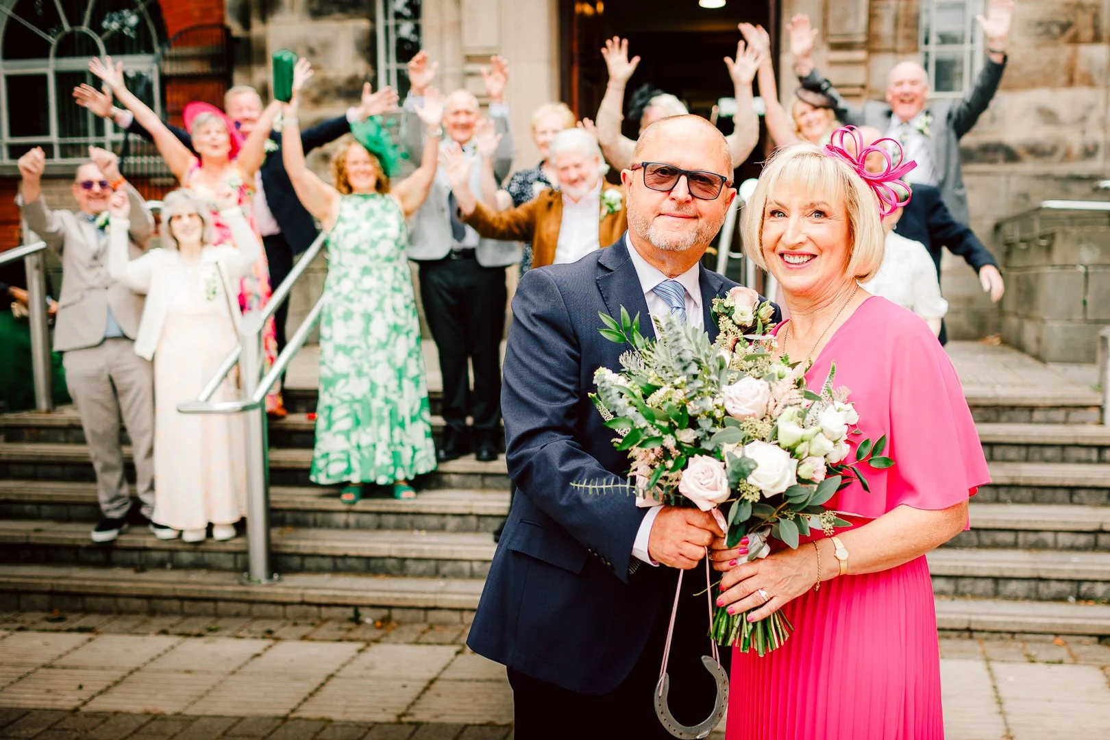 Happy newlyweds holding a floral bouquet on the steps with cheering guests behind them, in a photo created by a Sale Town Hall wedding photographer.