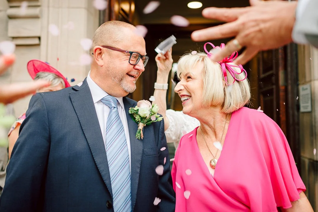 a couple step out of their wedding venue the bride dressed in luminous pink whilst the groom in a traditional blue suit. Celebrations go on around them with confetti visible