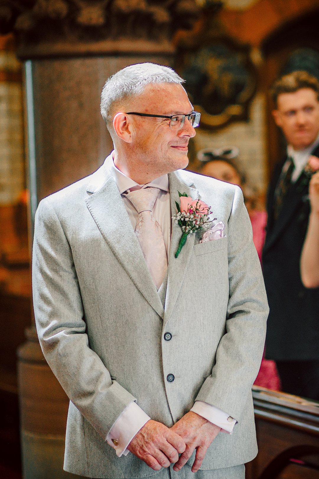 A middle-aged man in a light gray suit with a pink flower boutonniere, glasses, and a white dress shirt, standing indoors during a wedding ceremony.