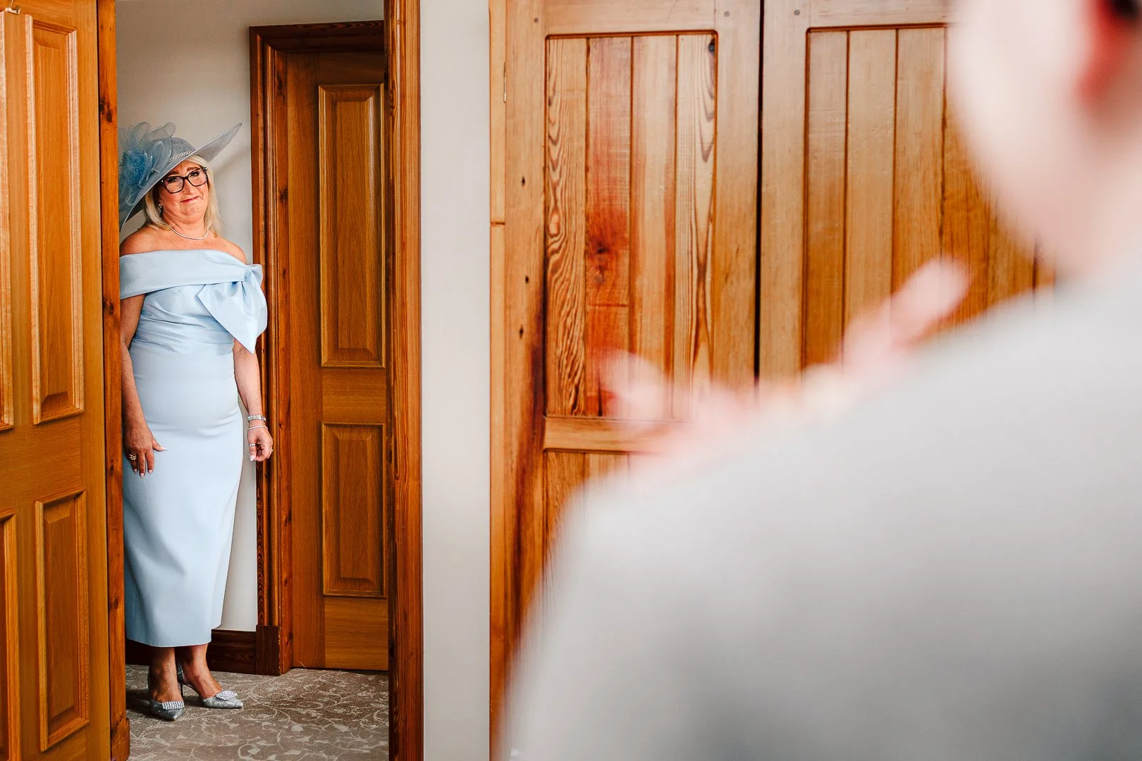 A woman dressed in a light blue gown with off-shoulder sleeves and a matching large hat with a veil, standing in a doorway, smiling at someone out of frame.