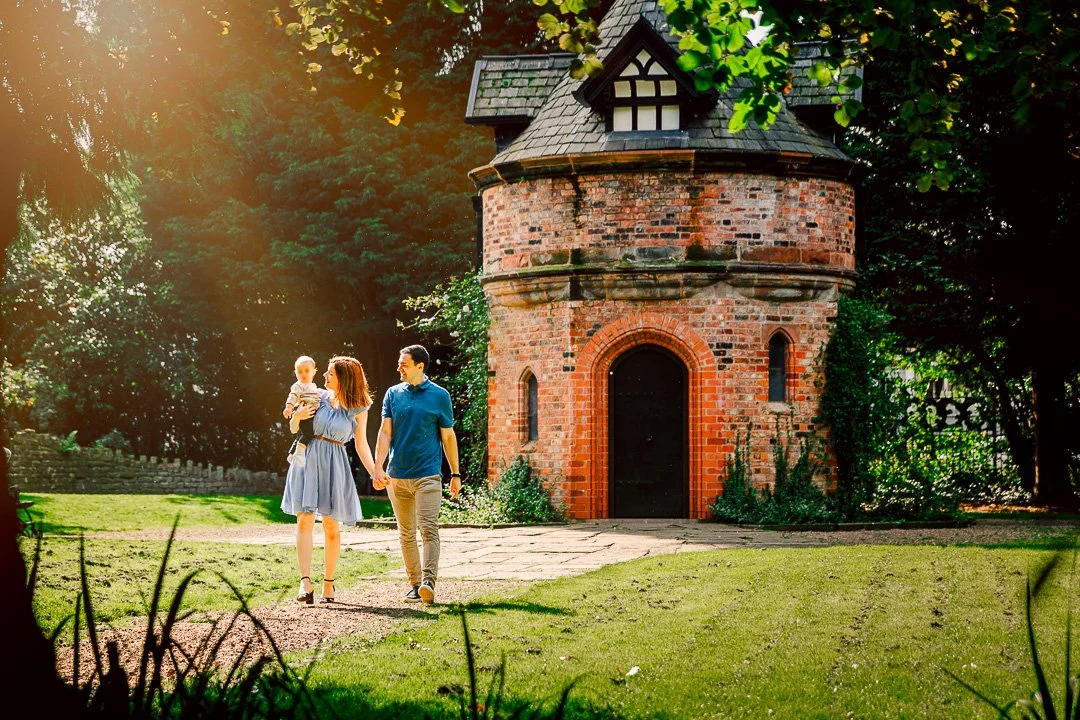 Parents walk holding baby in front of the Dovecot at Walkden Gardens in Sale during a Family Photoshoot