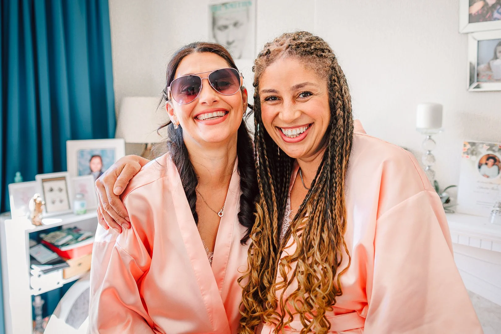 Two bridesmaids in pink robes smiling and embracing each other indoors during wedding prep in Sale