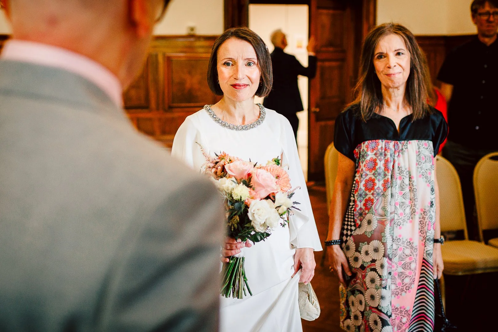 Bride walks aisle alongside her bridesmaid as they both look towards groom Dave in the ceremony room at Sale Town Hall