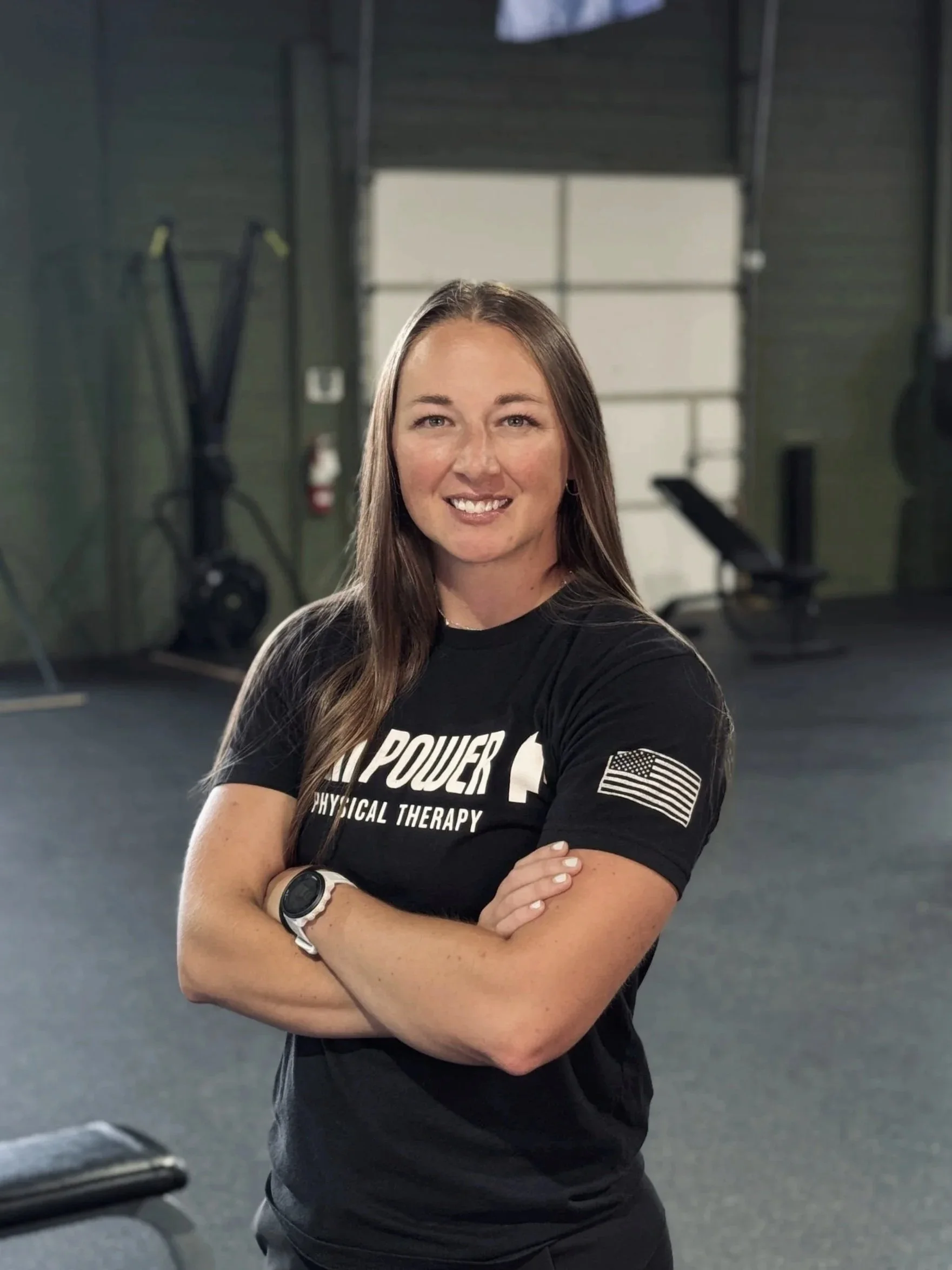A woman with long brown hair smiling, standing with crossed arms inside a gym, wearing a black T-shirt with white text and a covered logo, with gym equipment and a large window in the background.