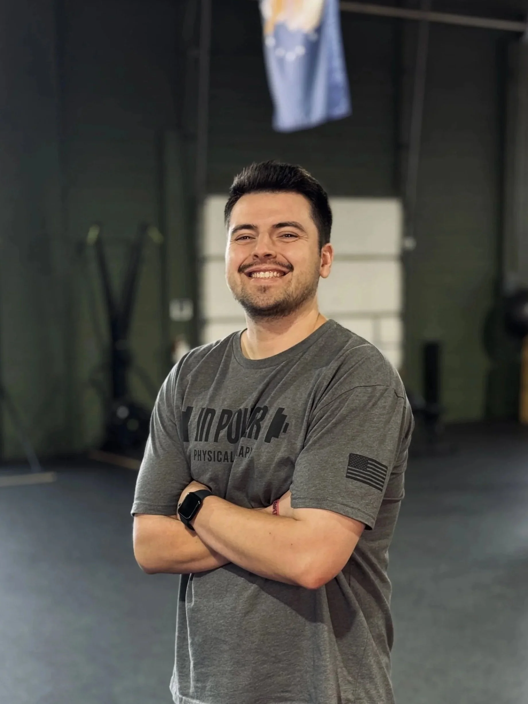 A smiling man with dark hair, wearing a gray T-shirt with black text, a black smartwatch, and a red wristband, standing with arms crossed in a gym or fitness studio with black walls and exercise equipment in the background.