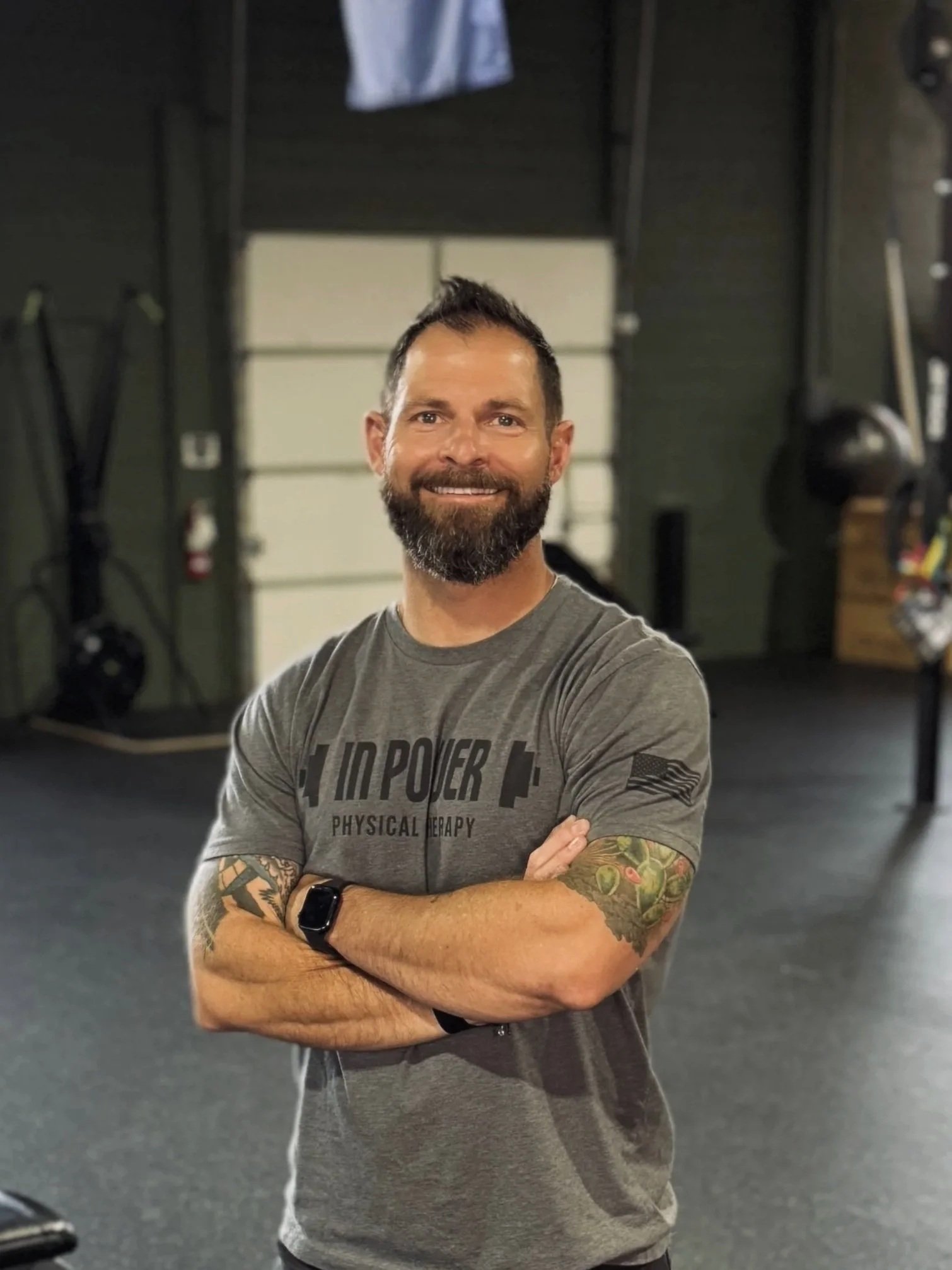A man with a beard and tattoos on his arms, wearing a gray T-shirt with printed text, is standing confidently with his arms crossed inside a gym. He has a broad smile and is wearing a smartwatch.