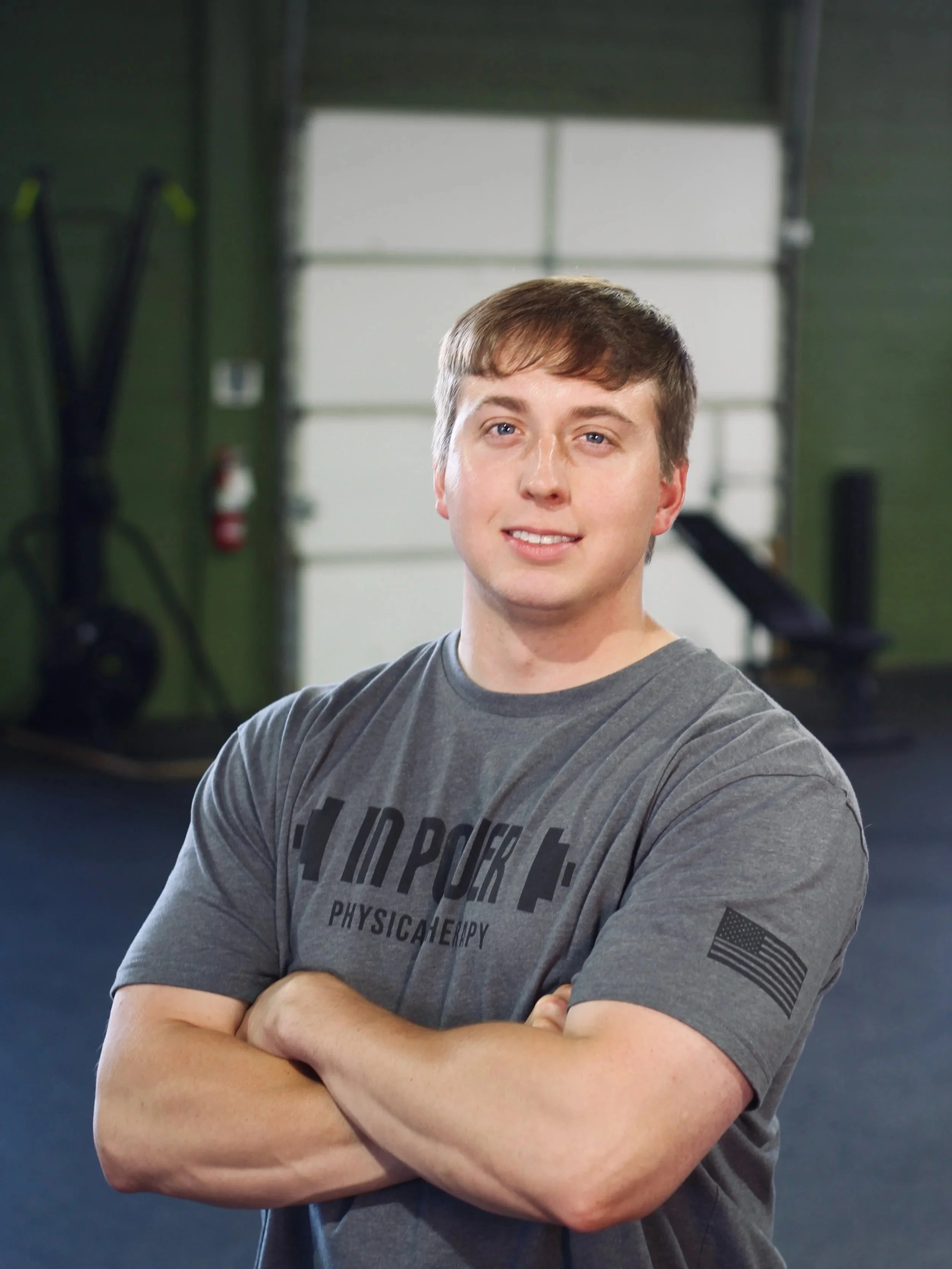 Young man standing with arms crossed, smiling, in a gym with fitness equipment and large windows in the background.