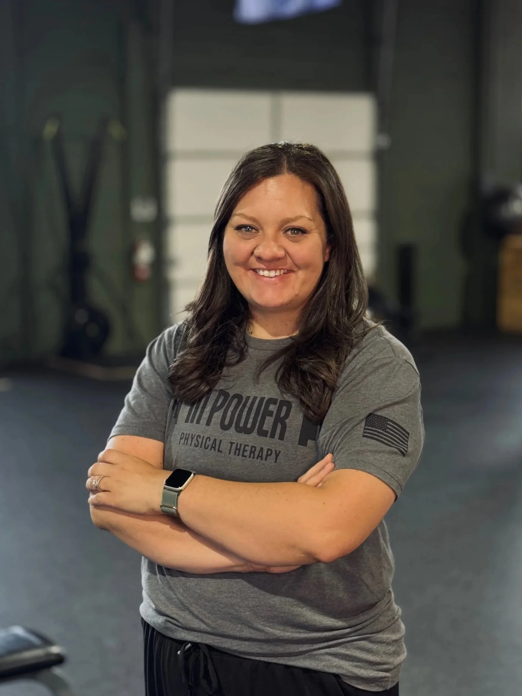 A smiling woman with long brown hair, wearing a gray t-shirt with black text, crossed arms, and a smartwatch on her left wrist, standing in a gym with a blurred background of gym equipment.
