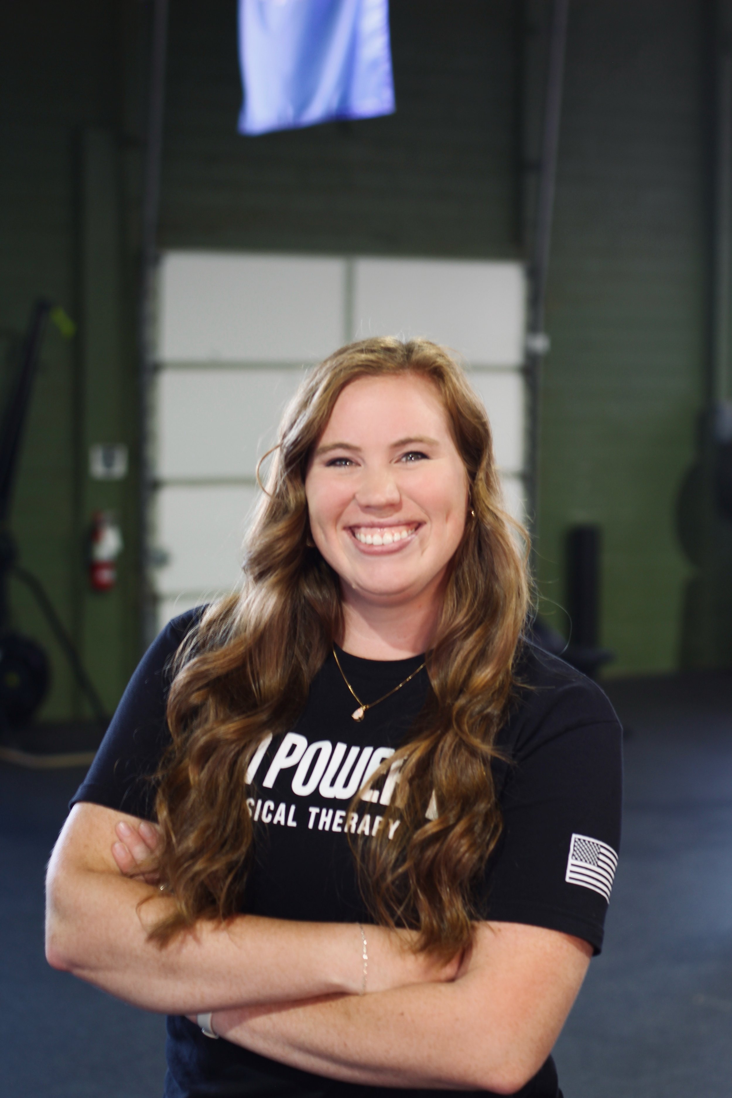 A young woman with long, wavy brown hair smiling with arms crossed, wearing a black shirt with partially visible white text, standing in an indoor gym or training facility.