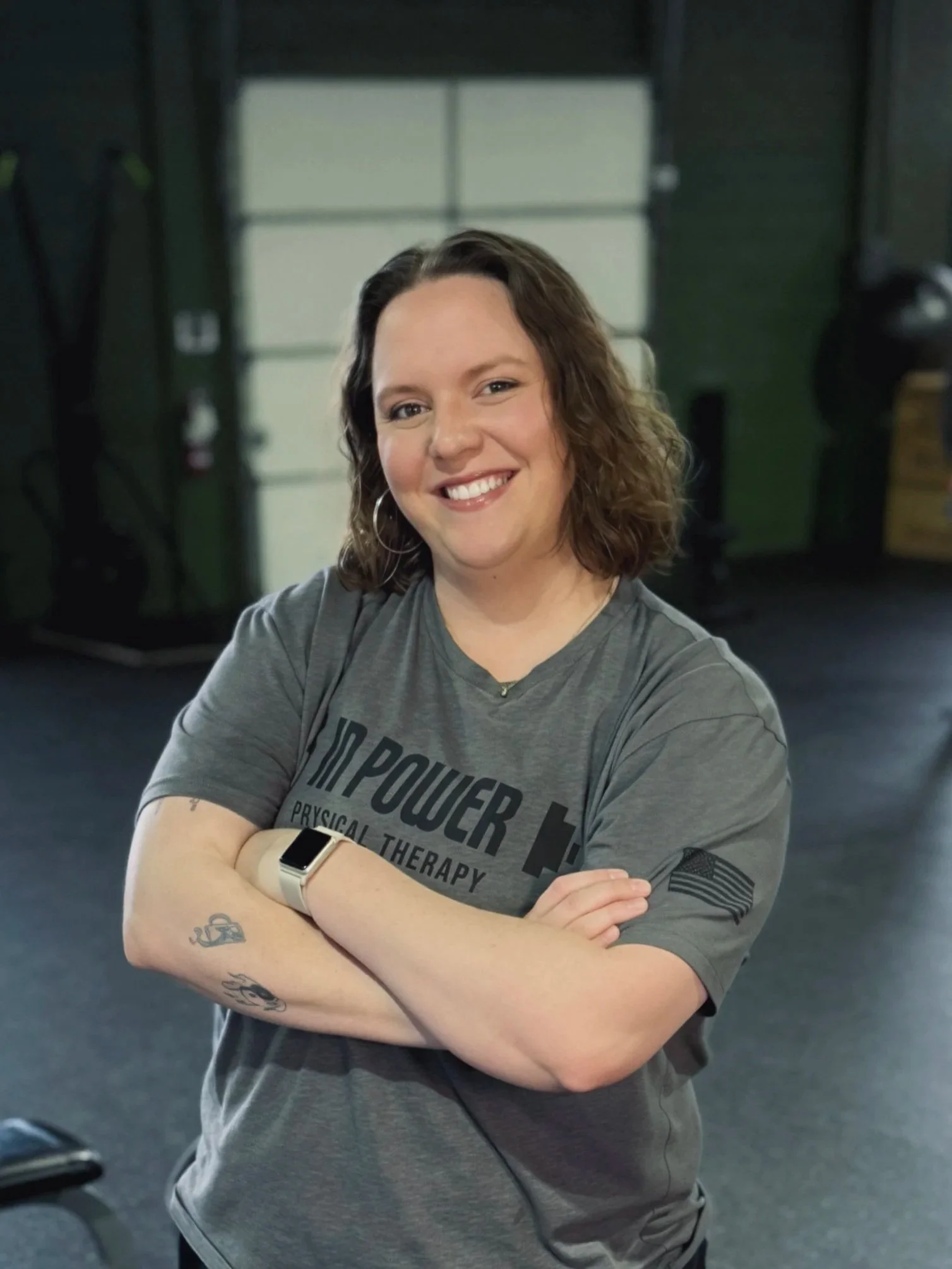 A woman with shoulder-length curly hair smiling with crossed arms wearing a gray t-shirt with military and fitness themes, standing inside a gym with fitness equipment in the background.