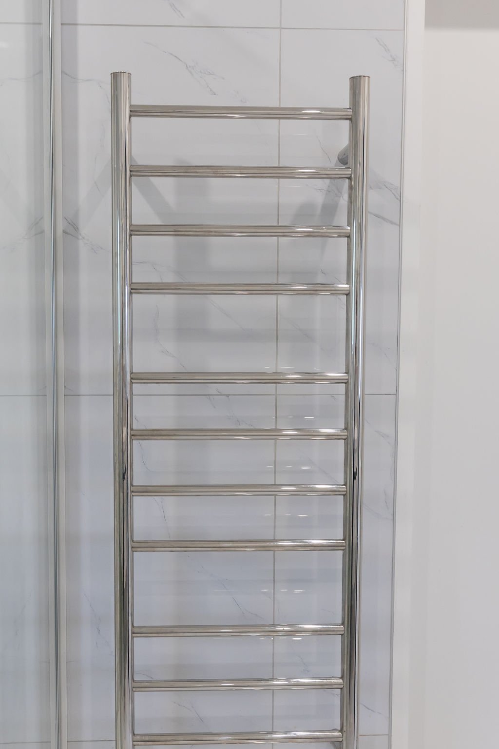 A silver heated towel rail with multiple horizontal bars mounted on a white tiled wall in a bathroom.