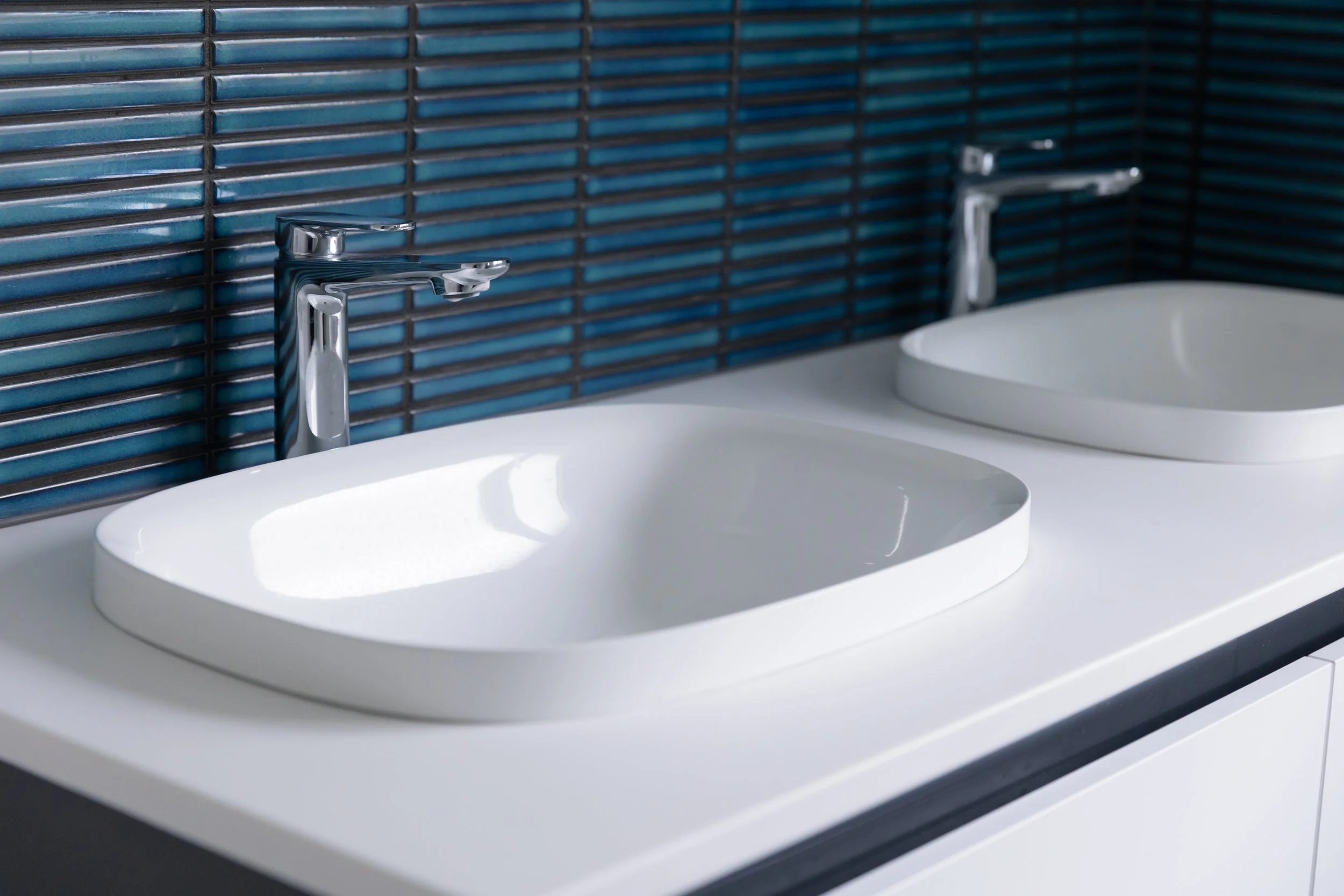 Two white bathroom sinks with chrome faucets in front of a blue tiled wall.
