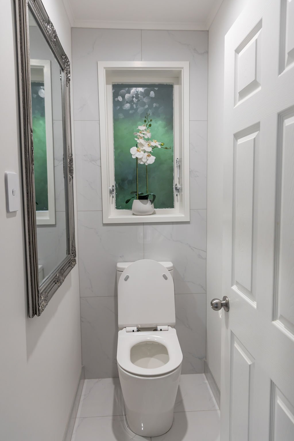 Small bathroom with white marble walls, a toilet, a window with an orchid plant, a large mirror with an ornate border, and a white door.