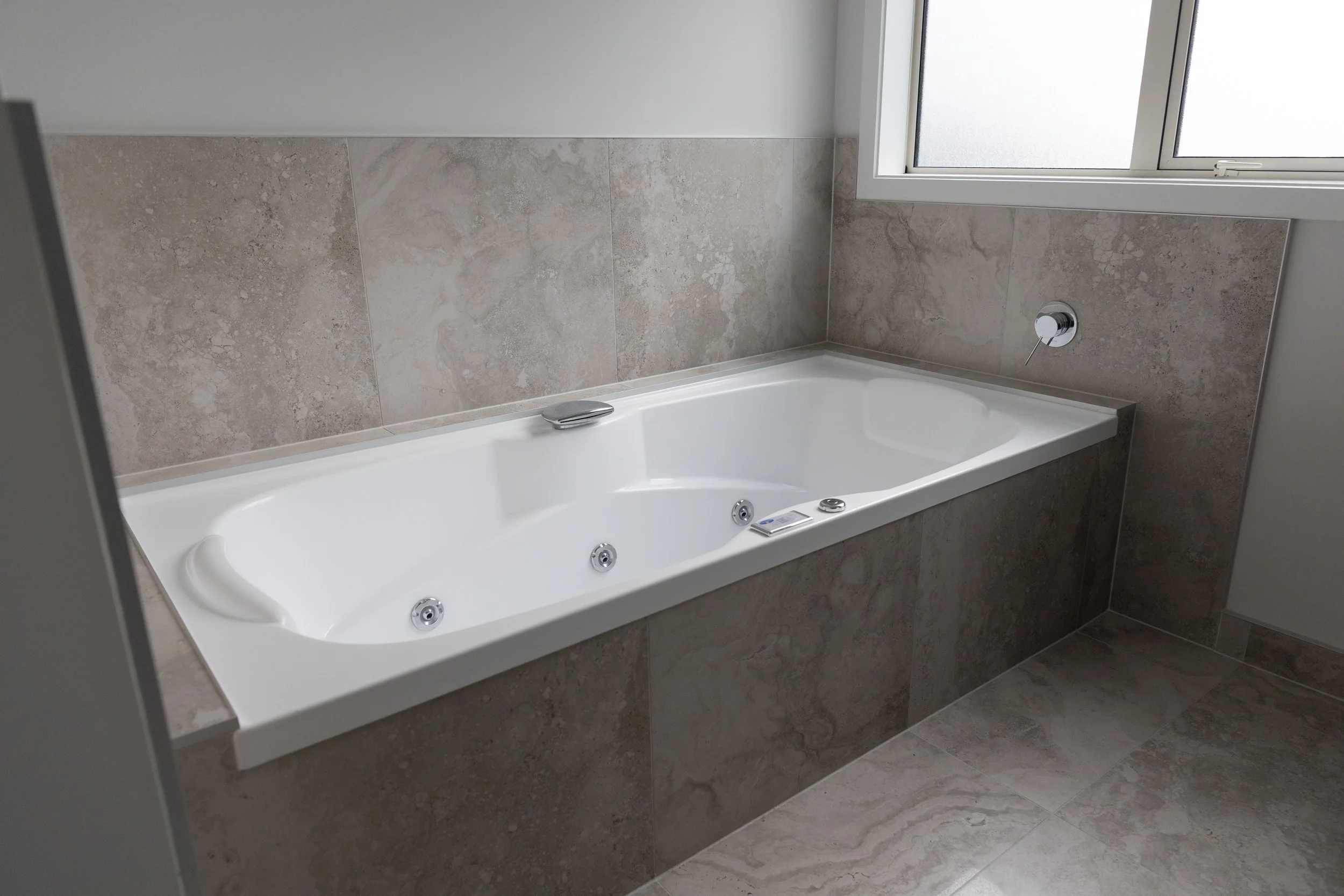 White bathtub with jets, installed in a bathroom with beige tiled walls and floor, near a frosted glass window.