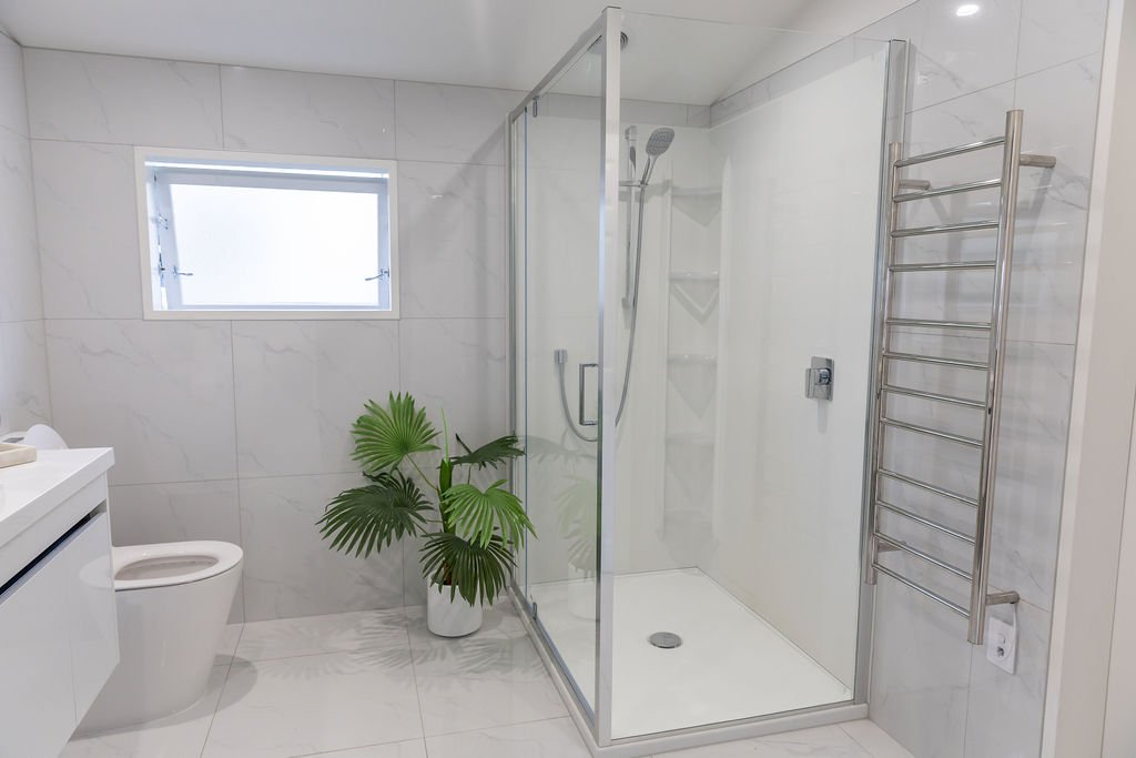 Modern bathroom featuring a glass-enclosed shower, a white toilet, a potted green plant, and a heated towel rack on a white tiled wall.