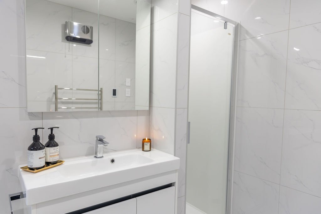 Modern bathroom with white marble walls, a white sink with two soap dispensers, a mirror, towel rack, small candle, and a glass shower enclosure.