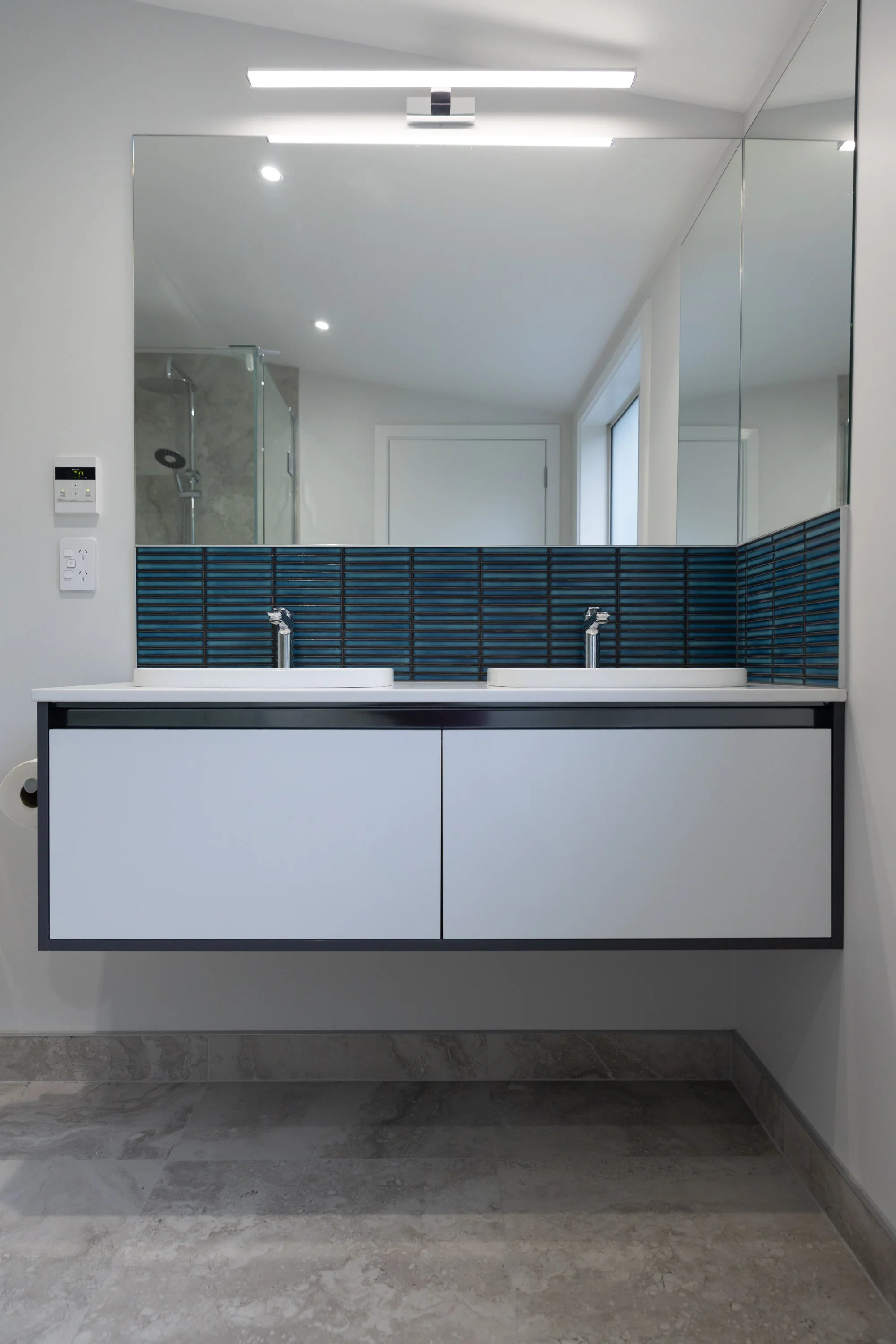 Modern bathroom with a large wall-mounted mirror, double sink vanity with blue tiled backsplash, and a walk-in shower in the background.