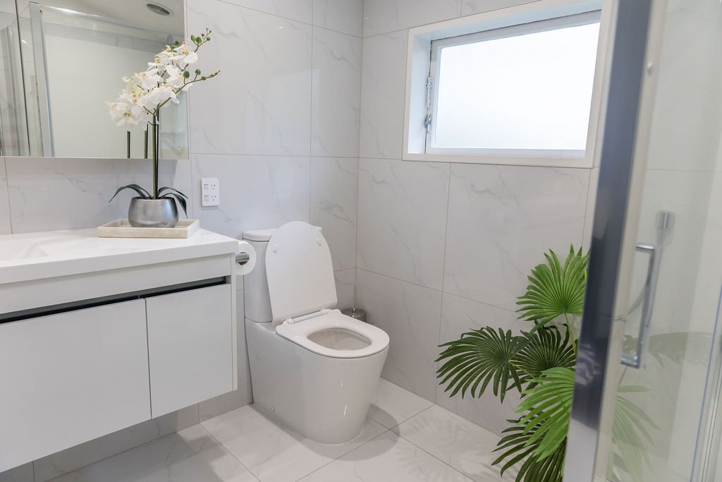 Modern bathroom with white marble tiles, a white toilet, a large green potted plant, a window, and a bathroom vanity with a mirror and a white orchid in a metallic pot.
