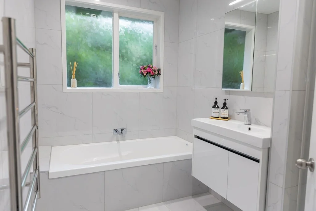 Modern bathroom with white marble walls, a bathtub under a frosted window, a small white sink with black handles, a large mirror above the sink, a metal heated towel rack on the left, and decorative items including a vase with pink flowers and two so