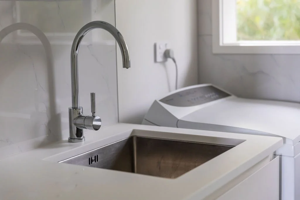 Laundry room with a stainless steel sink, chrome faucet, washing machine, and a window with view of greenery.