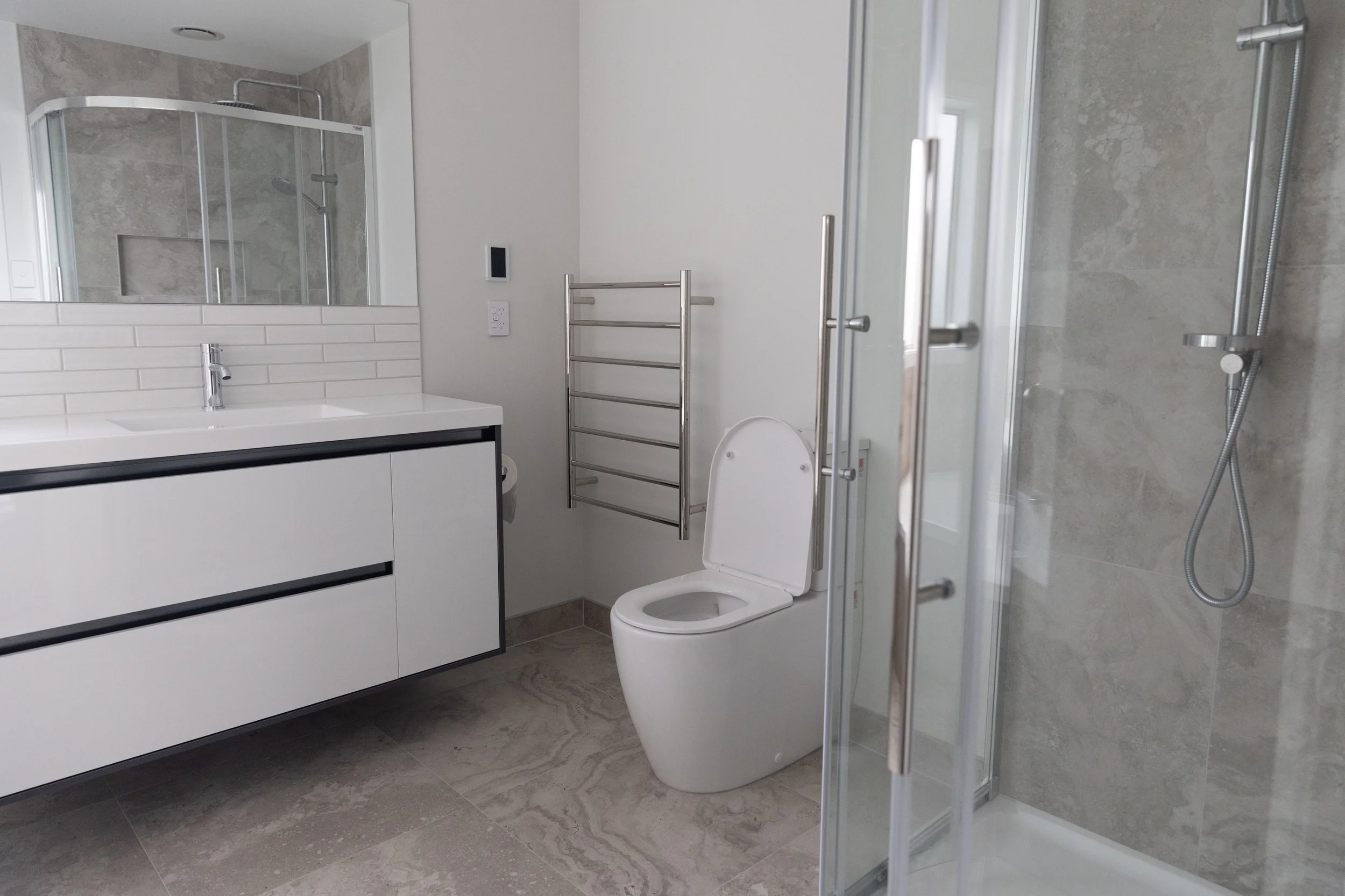 Modern bathroom with a white vanity, mirror, toilet, towel rack, and a glass-enclosed shower with gray tile walls.
