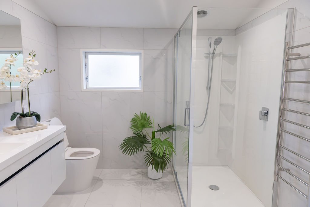 Modern bathroom with white tiled walls and floor, a frosted window, a white toilet, a potted plant, a vanity with orchid, and a glass-enclosed shower.