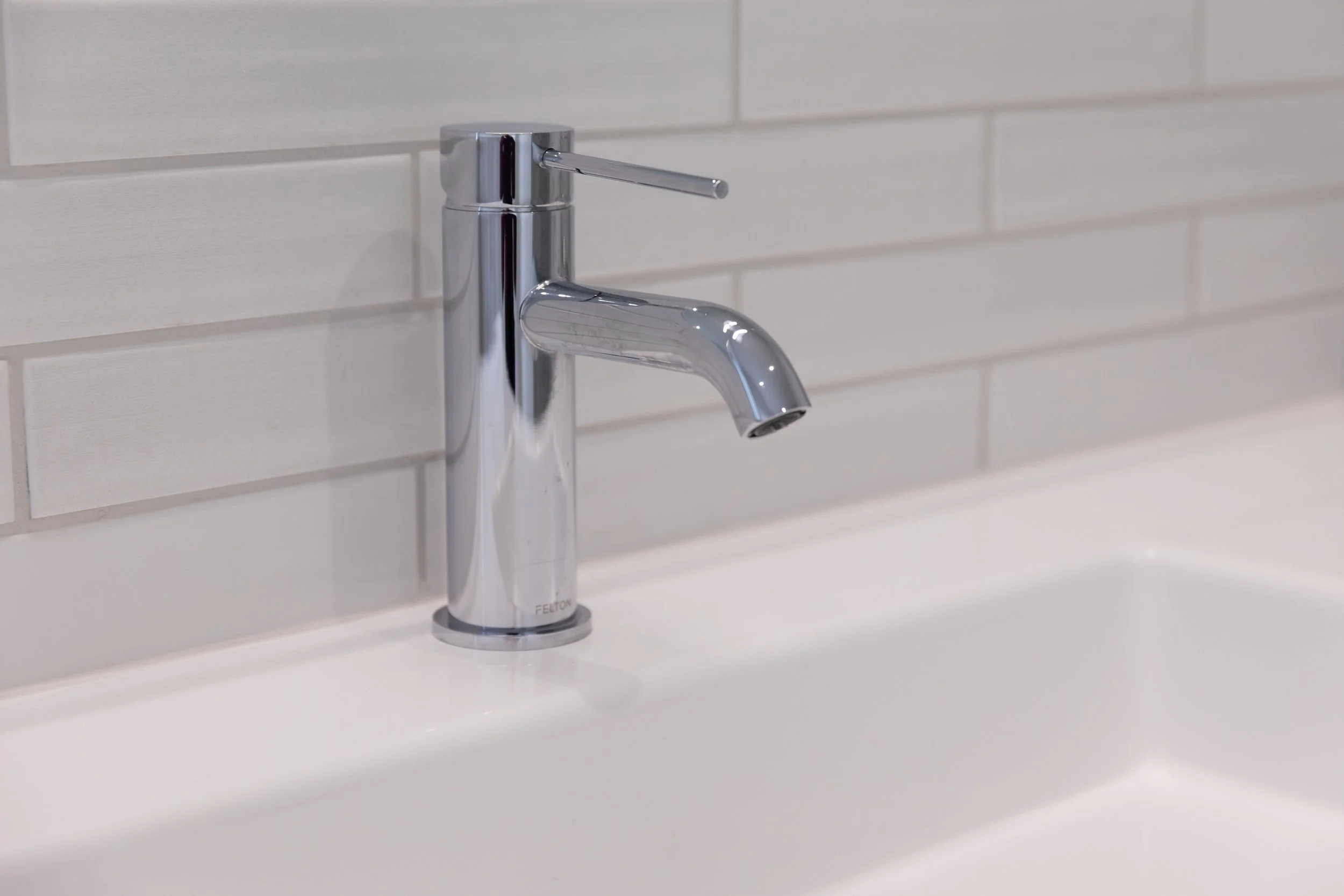 Chrome bathroom faucet mounted on white sink with light gray tiled wall in the background.