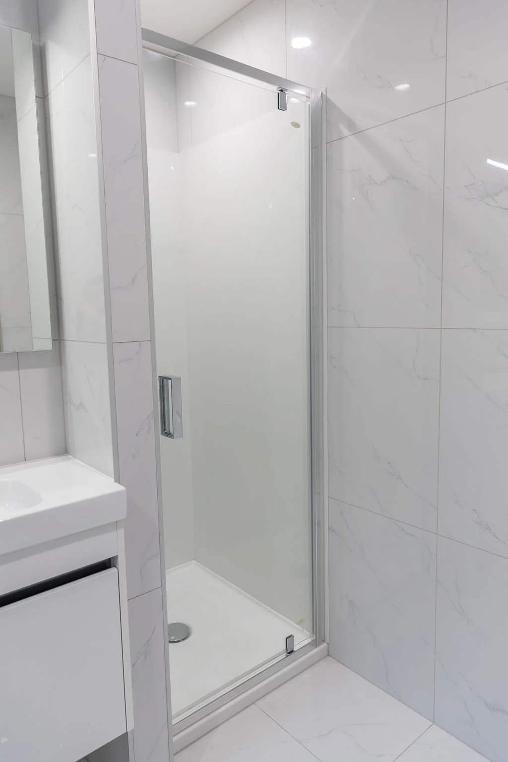 A modern bathroom shower with frosted glass door, white marble tiles on walls, and a small white vanity with a mirror.