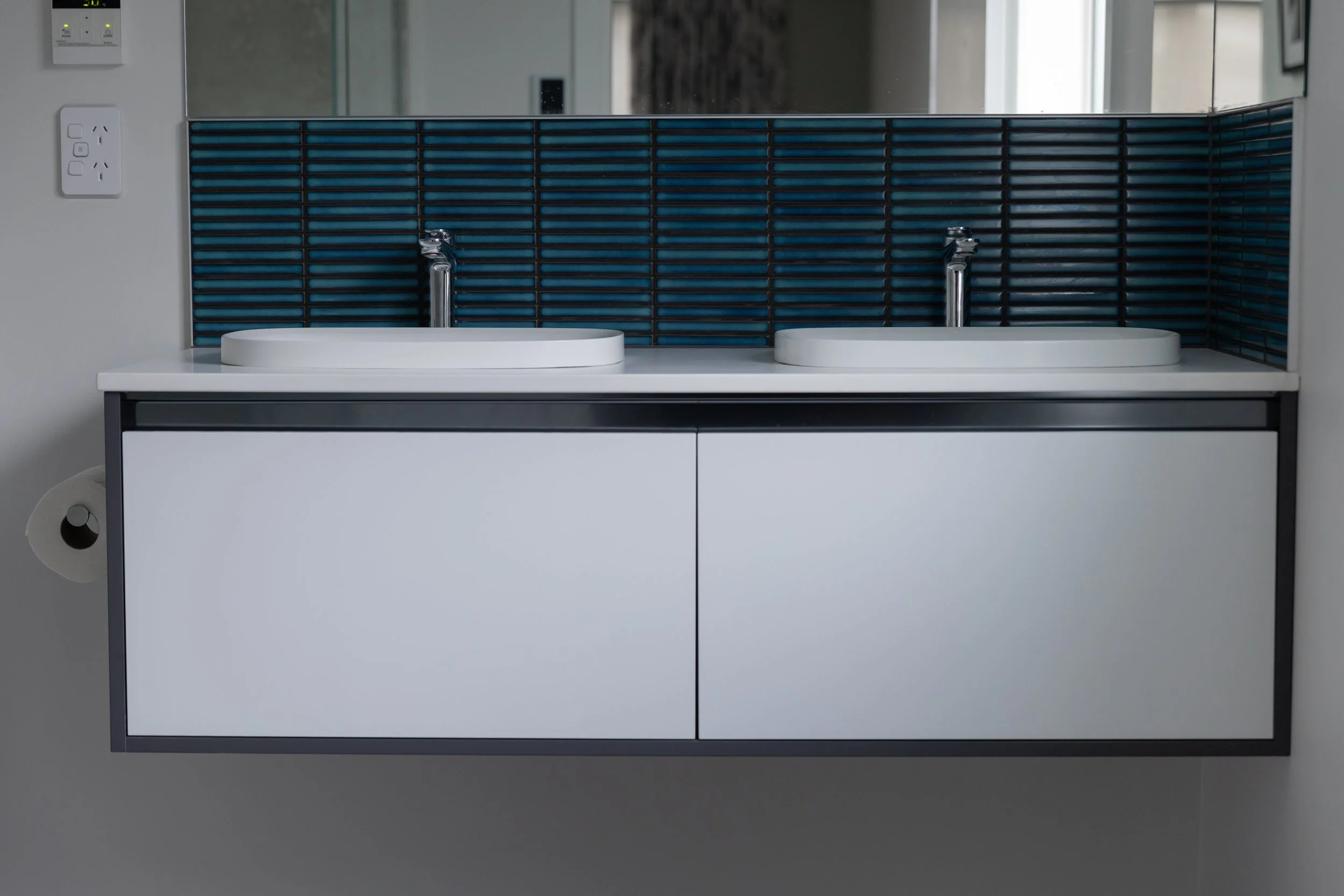 Modern bathroom vanity with two white sinks, chrome faucets, a black and blue tiled backsplash, mirrored wall, electrical outlets, and a toilet paper roll holder on the side.