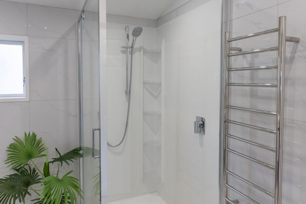Modern bathroom shower with a glass door, showerhead, corner shelves, towel radiator, window, and a potted green plant.
