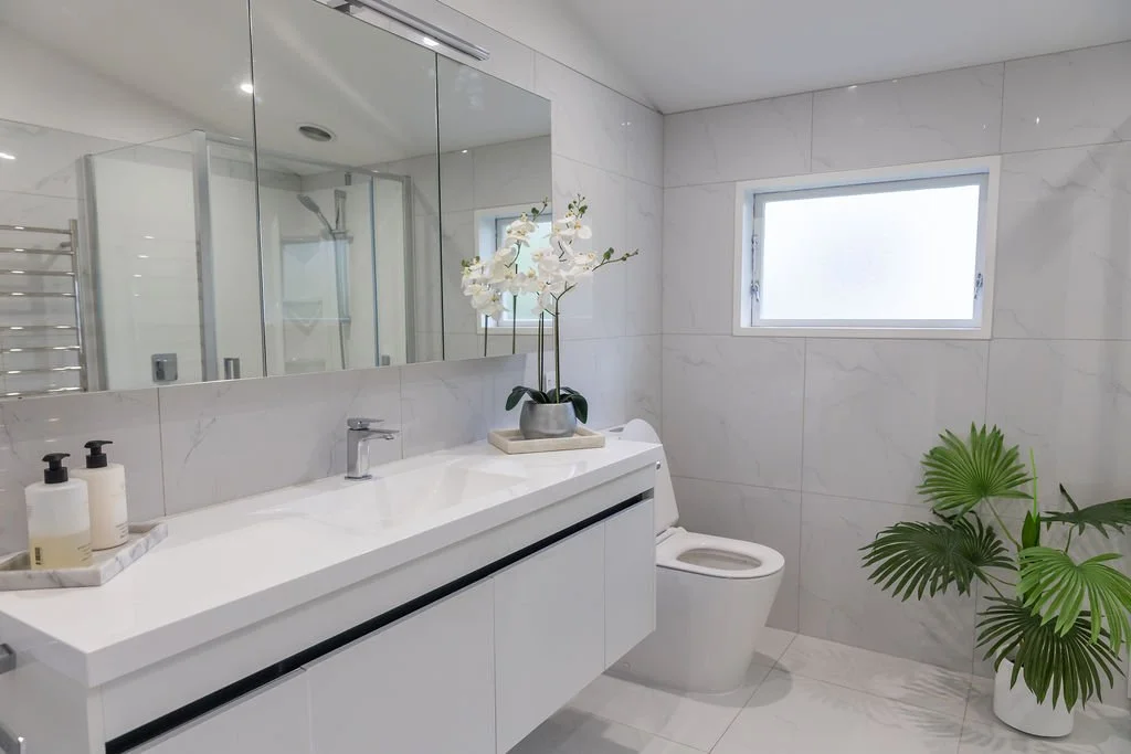 Modern white bathroom with marble walls, a large mirror, a white vanity with a sink, a toilet, a window, a potted plant, and a decorative orchid